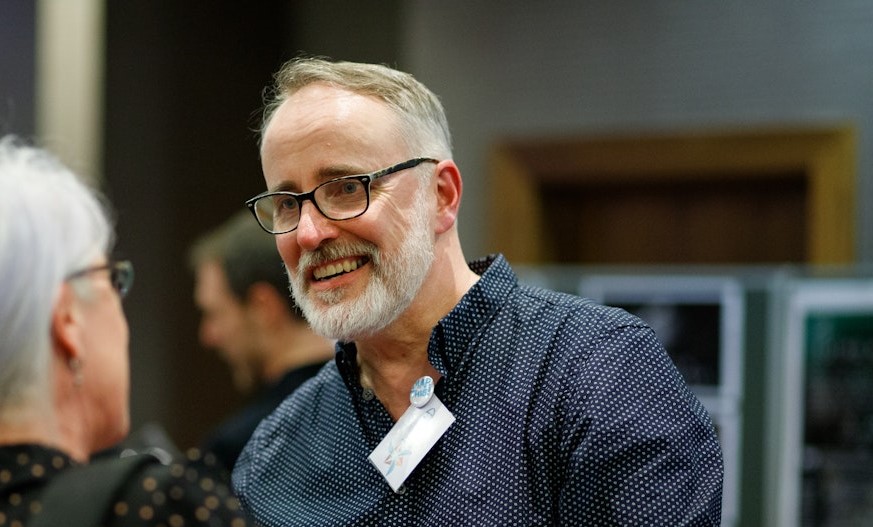 A man with short grey hair and beard with glasses talks to someone with their back to the camera.