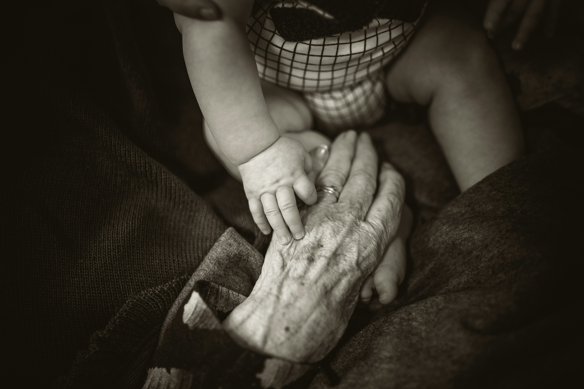 rod-long-y0OAmd_COUM-unsplash A baby's hand atop the hand of an older person. Black and white photograph.